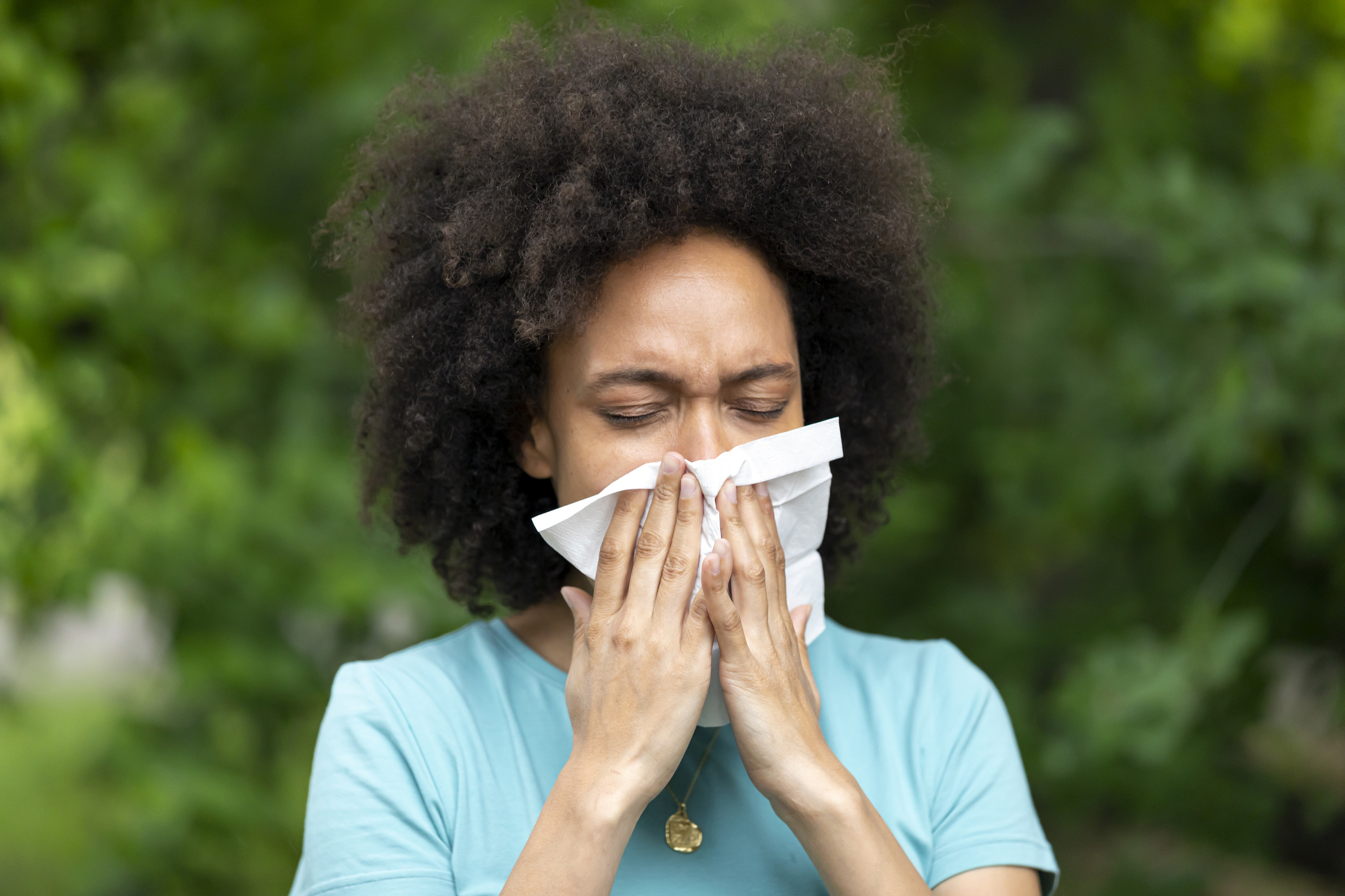 An African-American Woman with Sinusitis Problems is Feeling Displeased and Blowing Nose in Napkin During a Walk in City Park During a Summer Day.